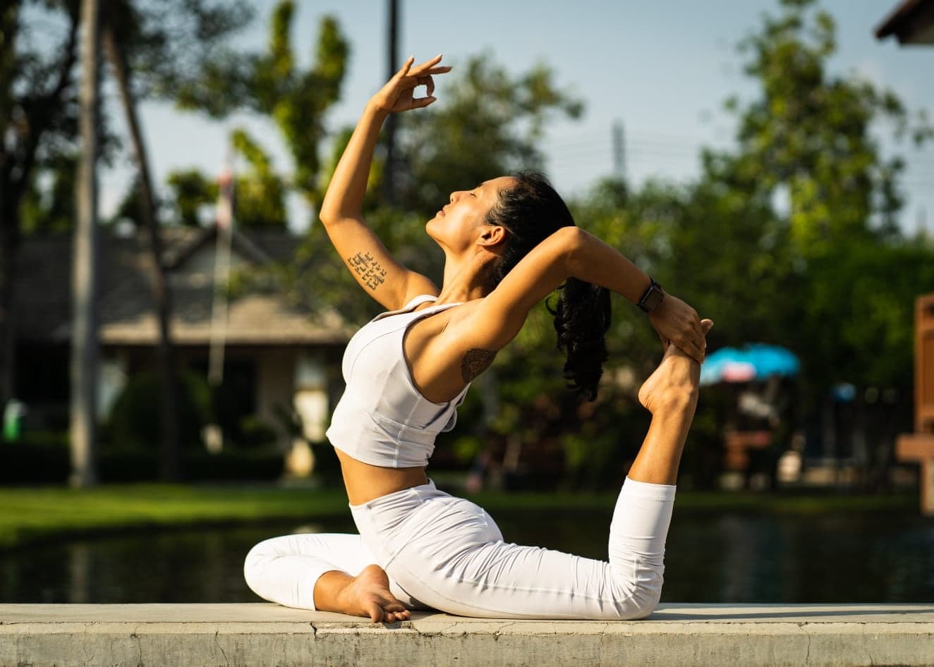 Nicha in a graceful seated backbend by the water, morning light on white clothing
